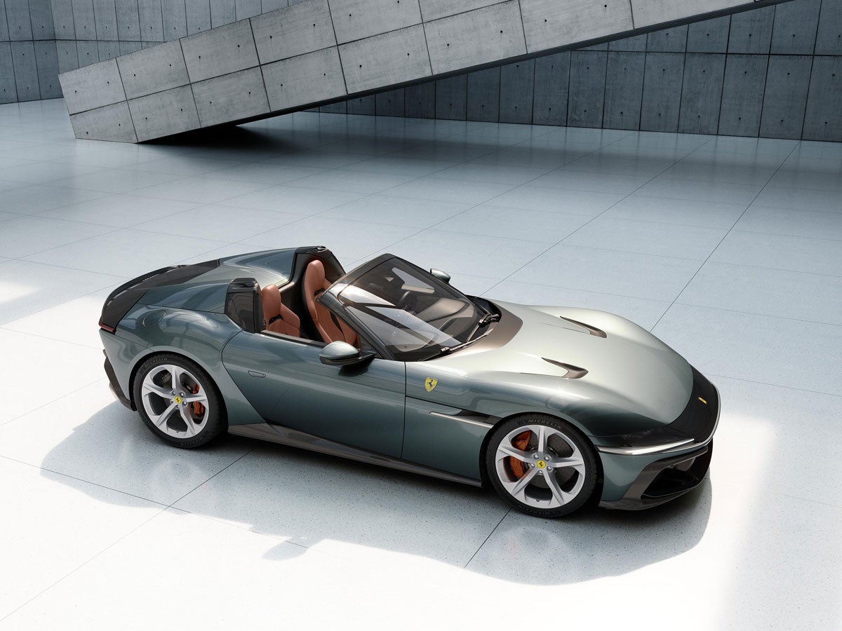 A teal Ferrari Monza SP2 convertible with tan leather interior is parked on a white tiled floor with an angled concrete structure in the background.