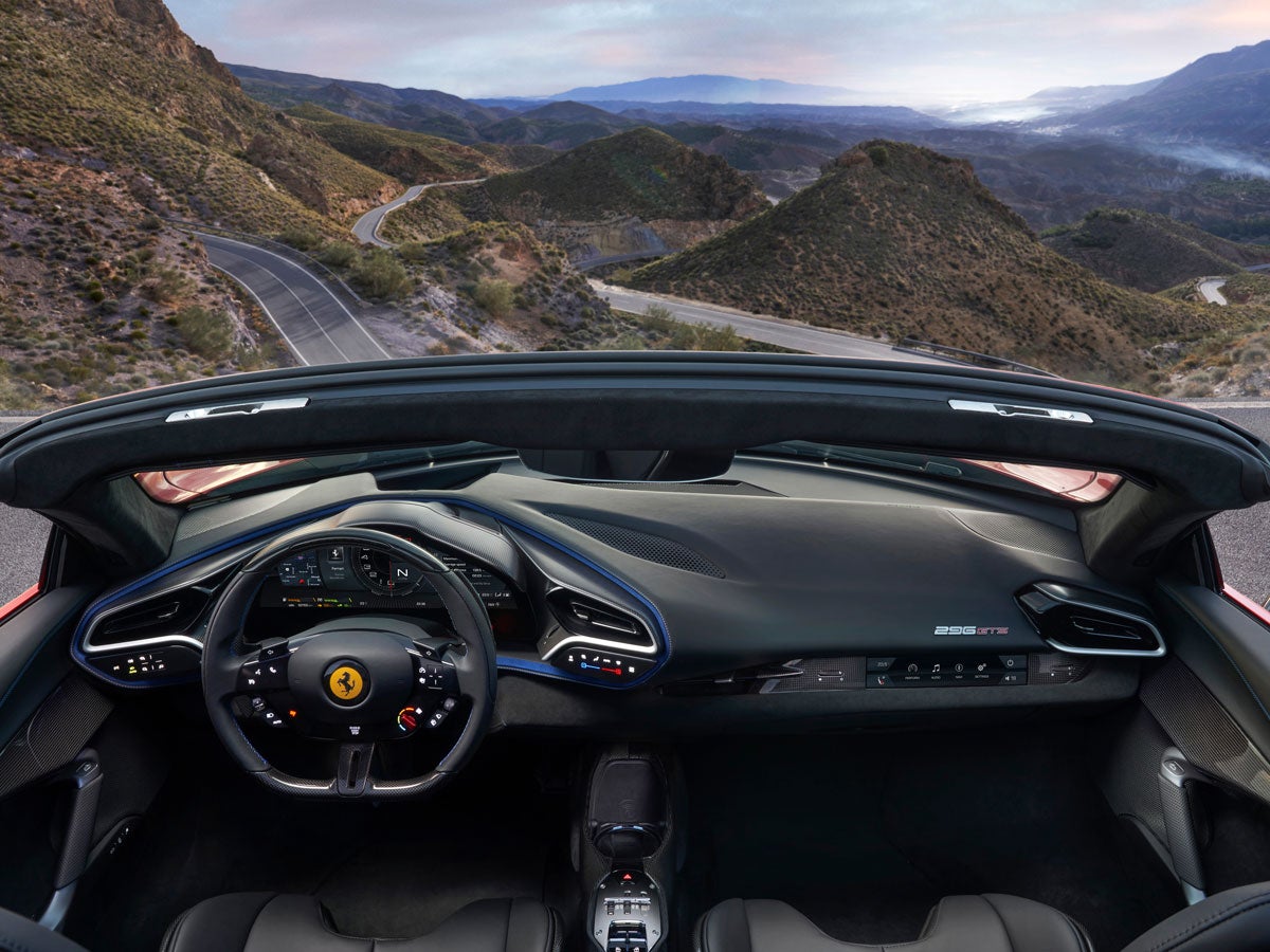 View from inside a convertible Ferrari 296 GTS, showing the dashboard, steering wheel, and a winding mountain road.