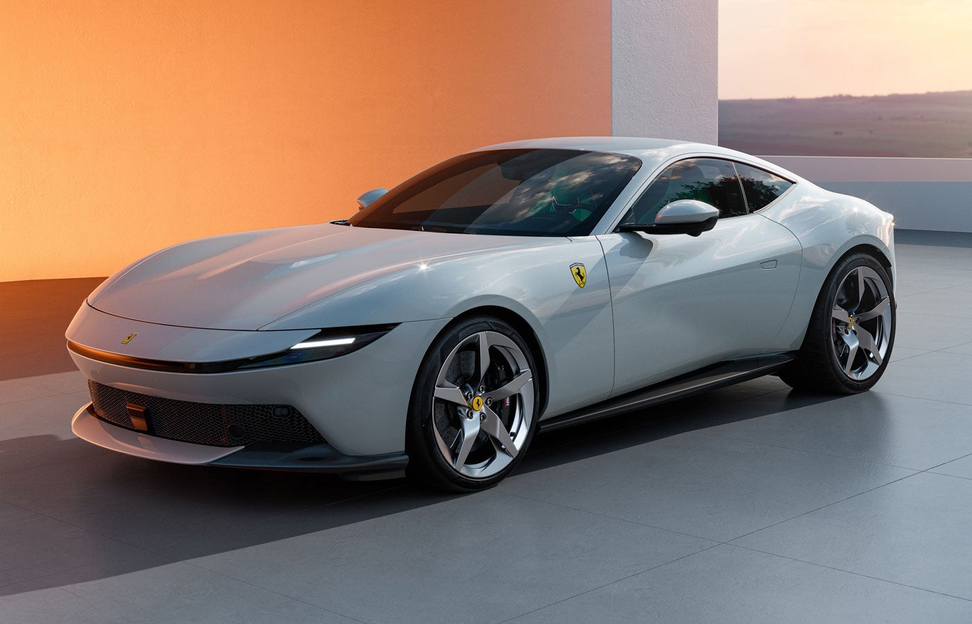 A light gray Ferrari, parked on a gray patio with an orange wall and distant landscape in the background.