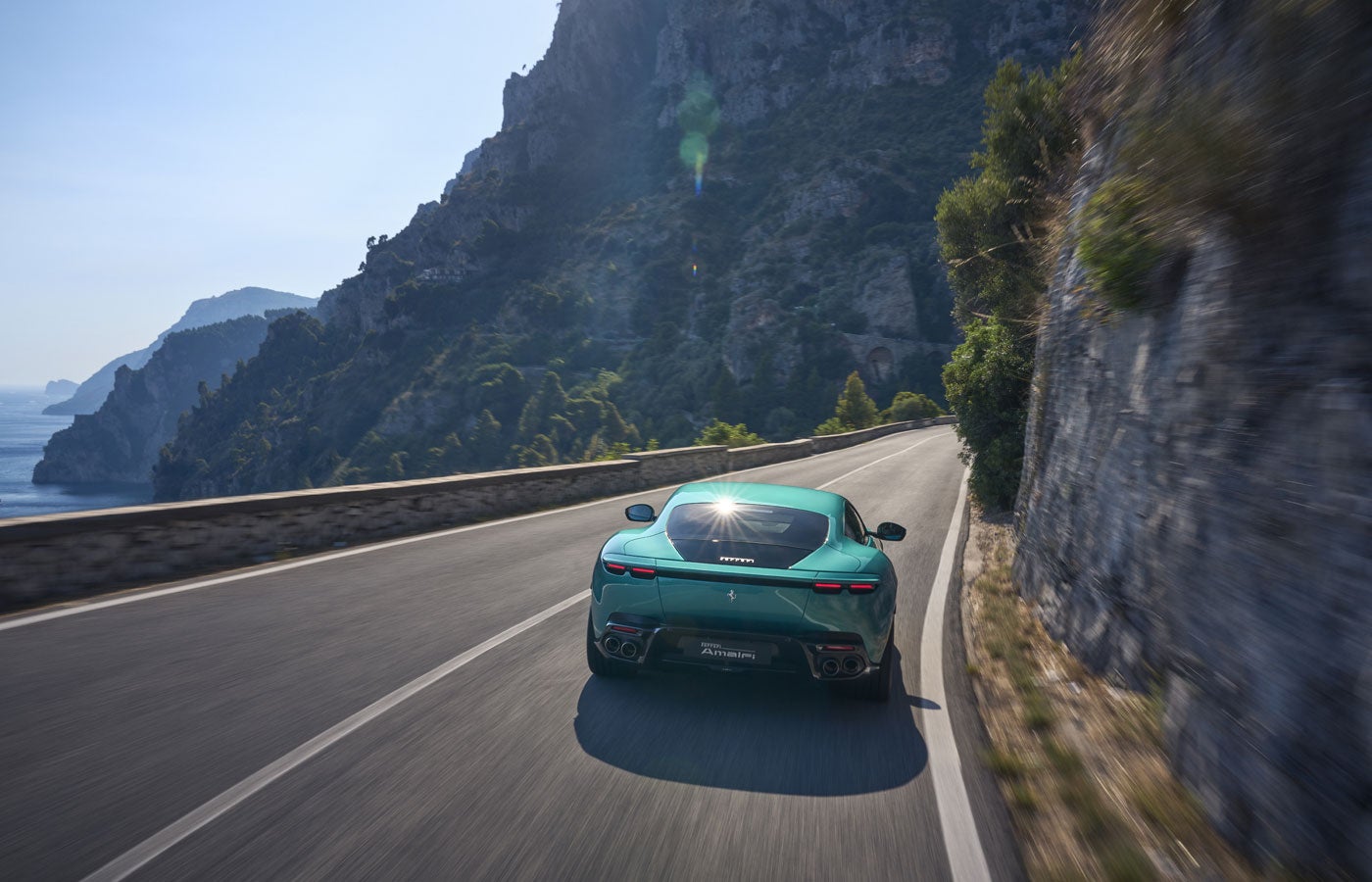 A light green Ferrari driving away on a scenic coastal road with mountains and the sea in the background.