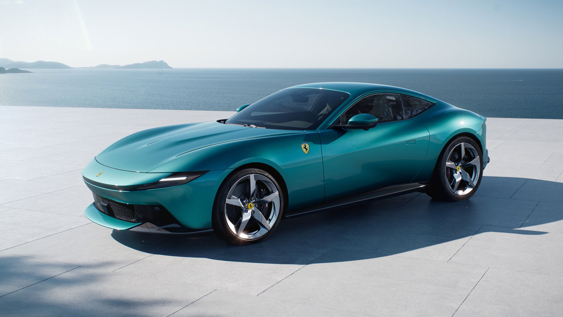 A teal Ferrari parked on a patio overlooking the ocean with distant mountains under a bright sky.
