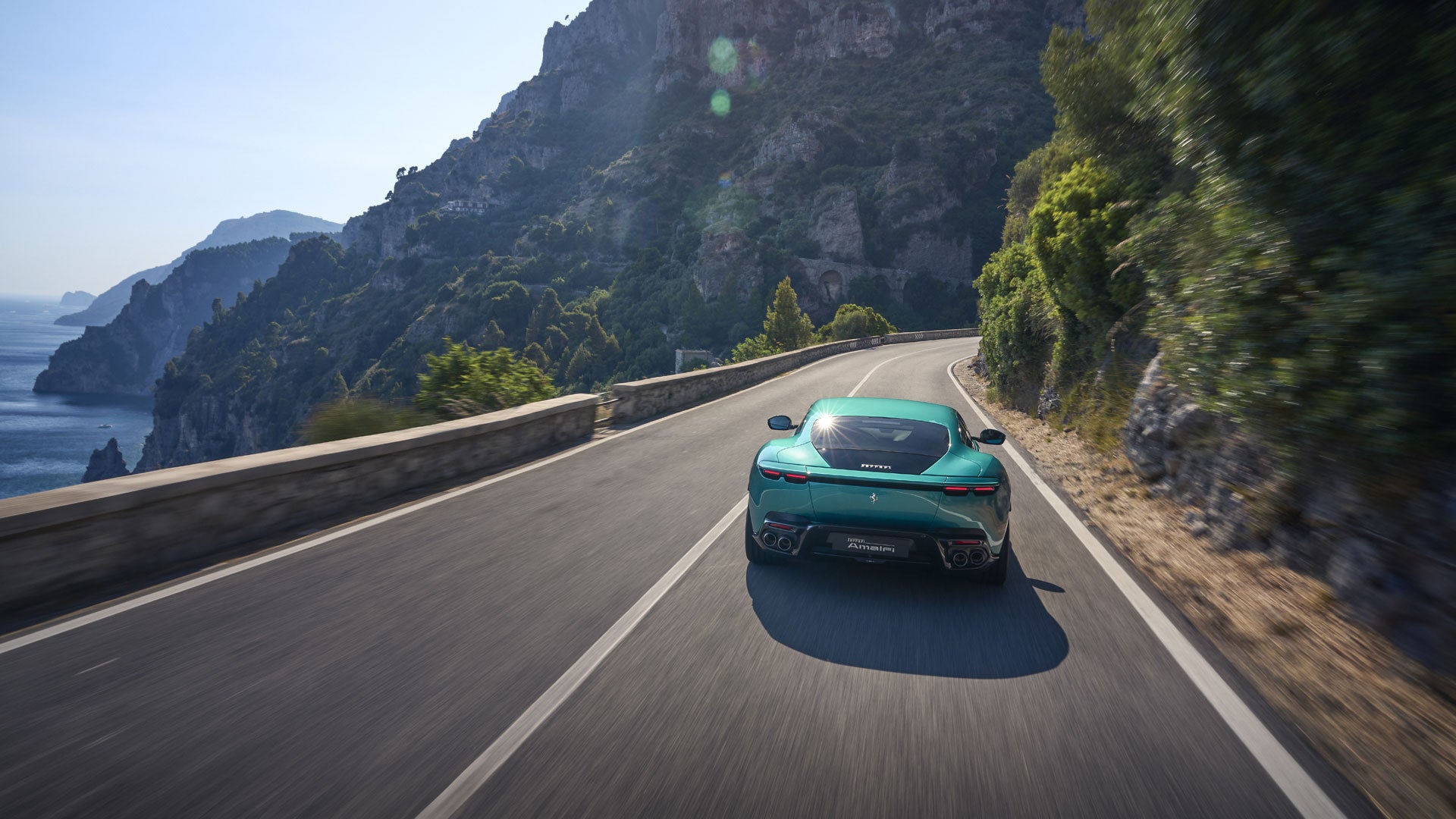 Teal Ferrari driving on a winding road next to an ocean with mountains in the background.